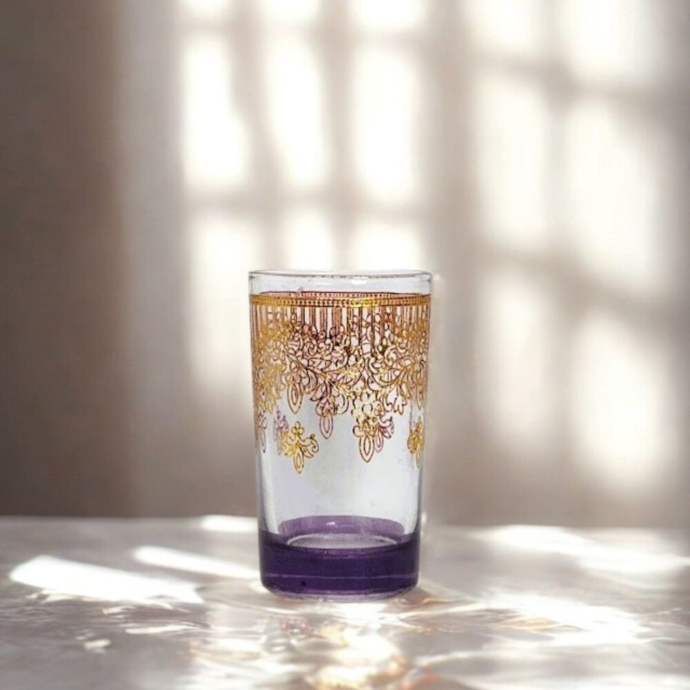 Elegantiška arbatos stiklinė su ryškiai violetiniu dugnu ir prabangiais aukso spalvos raštais, stovinti ant blizgaus paviršiaus. Fone matyti pro langą krentanti šviesa su lango rėmo šešėliais.