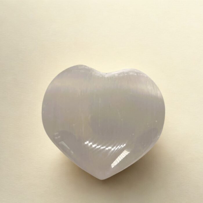Širdies formos poliruotas selenito akmuo, skirtas laikyti delne, gulintis ant švelnaus šviesaus audinio.