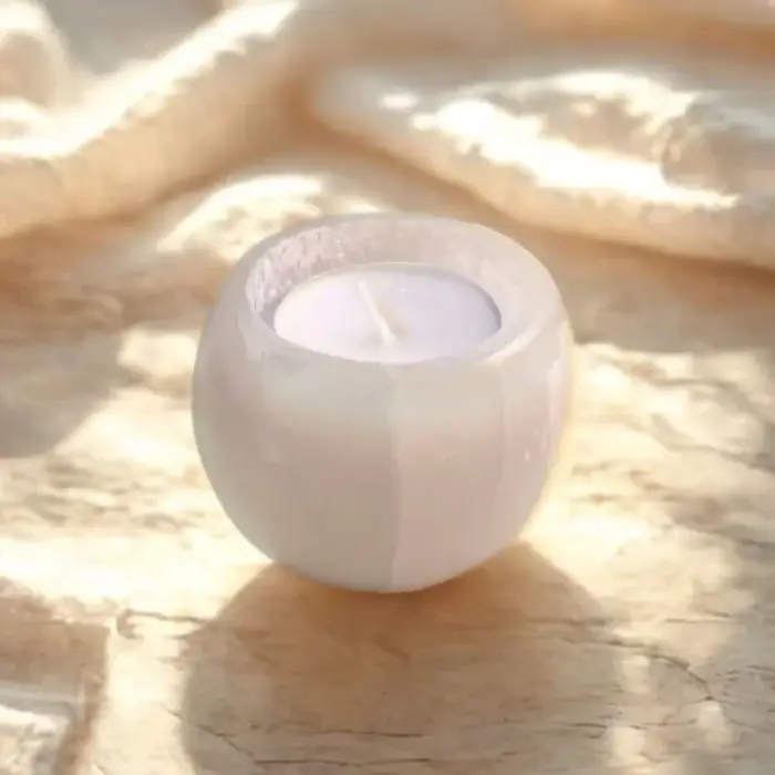 Nedidelė, apvali, balto selenito žvakidė „Mėnulis“ su arbatine žvake, stovinti ant gelsvo paviršiaus su šviesaus audinio klostėmis.