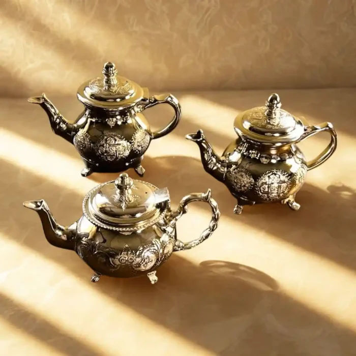 Trys rankų darbo sidabruoti arbatinukai, dekoruoti detaliais marokietiškais ornamentais, smėlio spalvos fone.