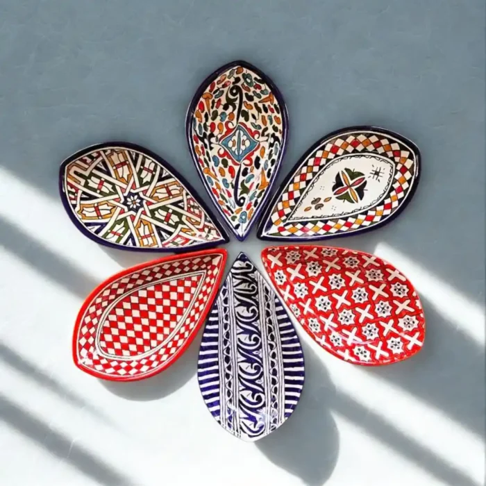 Šešios įvairiaspalvės, lašo formos keraminės lėkštės, išdėliotos apskritimu ant balto paviršiaus.