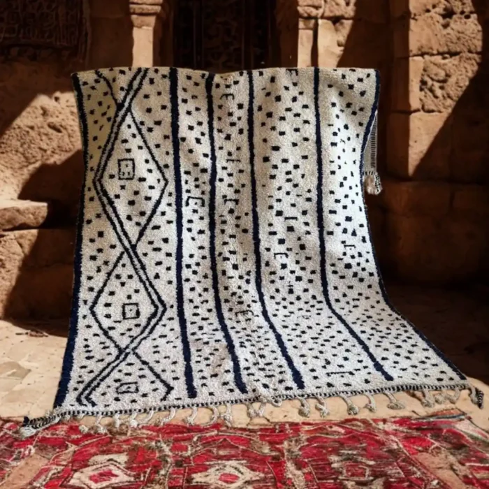 Berberų kilimas, baltas su tamsiai mėlynais geometriniais raštais, kabantis senovinio Maroko statinio fone.