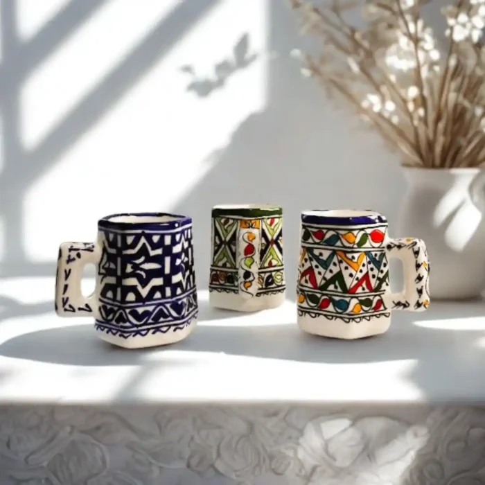 Trys keramikiniai marokietiški puodeliai su spalvotais raštais, išdėstyti ant baltos lentynos.