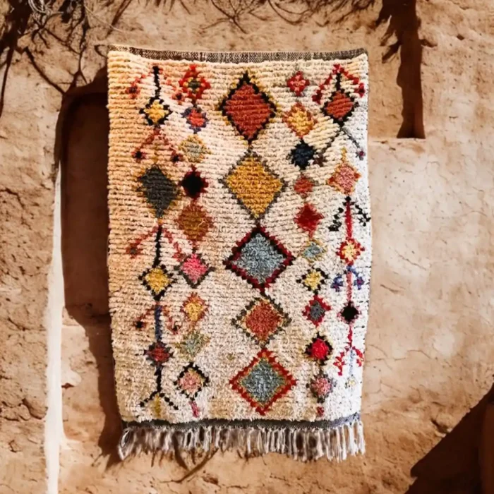 Tradicinis Maroko berberų kilimas su daugiaspalviais deimanto raštais ant kreminio fono, pakabintas ant senovinės molio sienos.